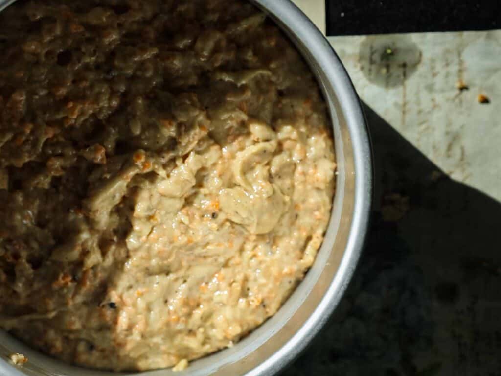 A close-up of a vegan carrot cake batter in round cake pan. The mixture appears to have a light brown color with small dark flecks throughout and is placed on a surface with shadows visible in the background.